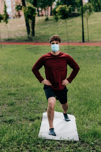 Joven deportista en máscara médica con las manos en las caderas haciendo ejercicio en la esterilla de fitness en el parque - foto de stock