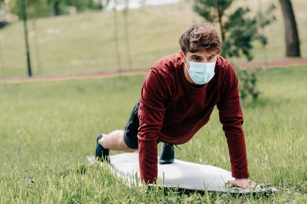 Deportista en máscara médica haciendo flexiones durante el ejercicio en la estera de fitness en la hierba en el parque - foto de stock