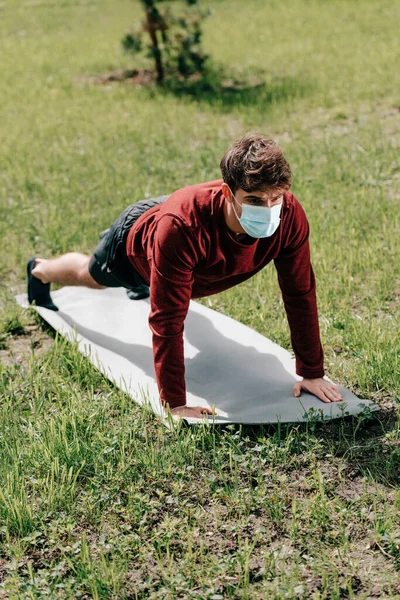 Спортсмен в медицинской маске делает отжимания во время тренировки на фитнес-коврик в парке — стоковое фото