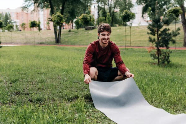 Bonito homem sorrindo ao colocar tapete de fitness na grama no parque — Fotografia de Stock