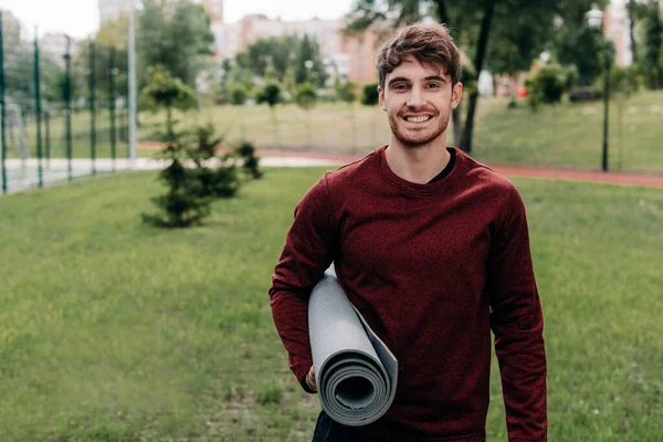 Handsome sportsman smiling at camera while holding fitness mat in park — Stock Photo