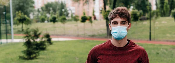 Foto panoramica dell'uomo in maschera medica che guarda la macchina fotografica nel parco — Foto stock