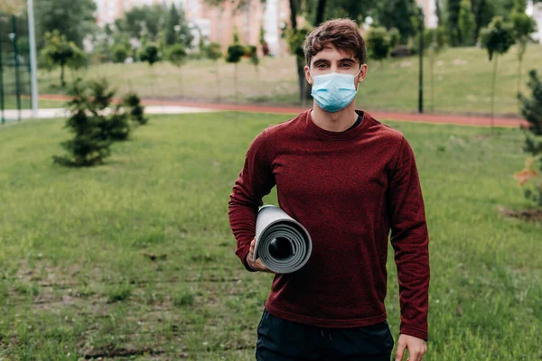 Человек в медицинской маске держит фитнес-мат, стоя в парке — стоковое фото