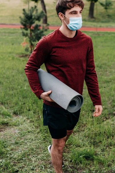 Deportista en máscara médica sujetando la alfombra de fitness mientras camina en el parque - foto de stock