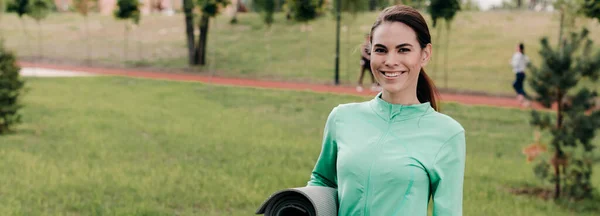 Orientação panorâmica da bela esportista sorrindo para a câmera enquanto segura o tapete de fitness no parque — Fotografia de Stock