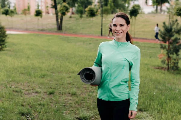 Attractive sportswoman holding fitness mat while smiling at camera in park — Stock Photo