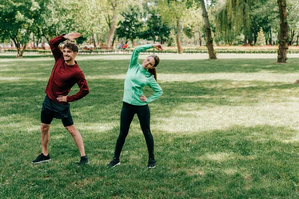 Hombre guapo sonriendo mientras hace ejercicio cerca de hermosa novia en el parque - foto de stock