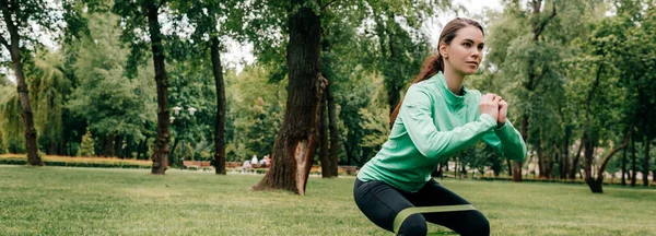 Panoramic crop of sportswoman doing squat with resistance band in park — Stock Photo