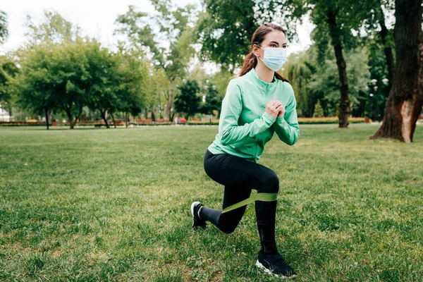 Young sportswoman in medical mask training with resistance band in park — Stock Photo