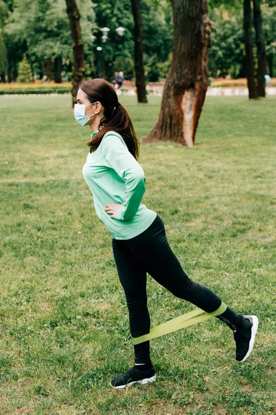 Vista laterale della sportiva in maschera medica utilizzando la fascia di resistenza durante l'allenamento nel parco — Foto stock