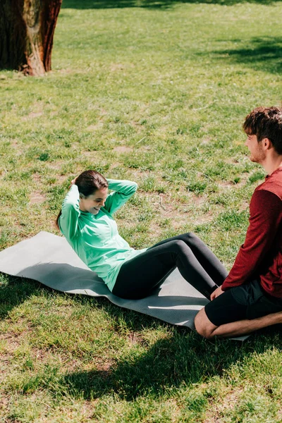 Esportista sorrindo fazendo abdominais perto de namorado no tapete de fitness no parque — Fotografia de Stock