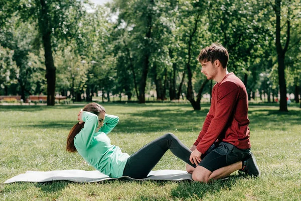 Vista lateral del hombre apoyando a la deportista mientras hace ejercicio en la estera de fitness en el parque - foto de stock