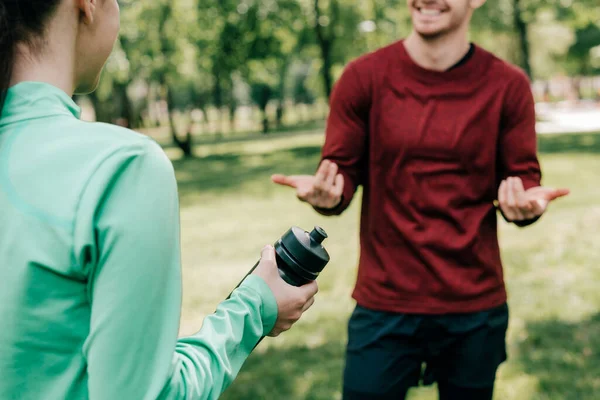 Vista cortada de esportista segurando garrafa de esportes perto de namorado sorridente no parque — Fotografia de Stock