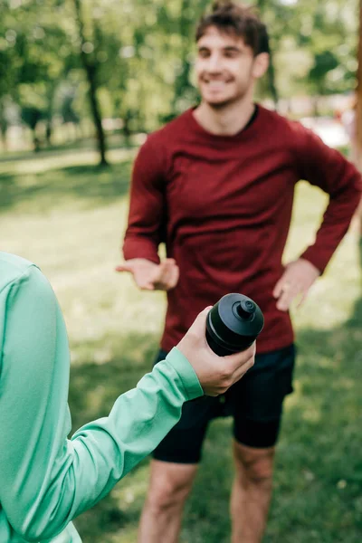 Foco seletivo de esportista segurando garrafa de esportes perto de sorrir desportista no parque — Fotografia de Stock