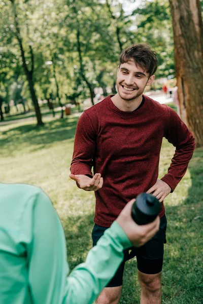 Enfoque selectivo de deportista guapo hablando con novia con botella de deporte en el parque - foto de stock