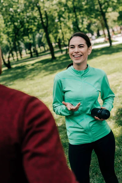 Selective focus of smiling sportswoman talking to boyfriend while holding sports bottle in park — Stock Photo