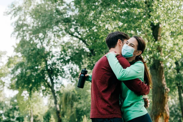 Desportista abraçando namorada em máscara médica segurando garrafa de esportes no parque — Fotografia de Stock