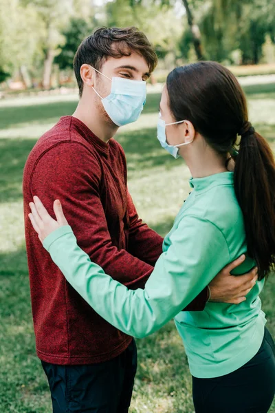 Vista laterale della sportiva in maschera medica abbracciando fidanzato nel parco — Foto stock