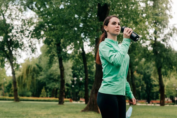 Joven deportista sosteniendo máscara médica y botella deportiva en el parque - foto de stock