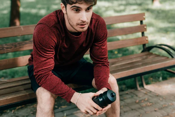 Красивый спортсмен держит спортивную бутылку, сидя на скамейке в парке — стоковое фото