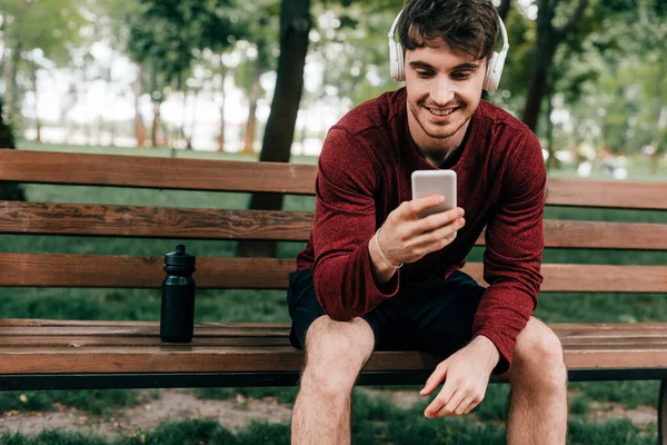 Esportista sorridente em fones de ouvido usando smartphone perto de garrafa de esportes no banco no parque — Fotografia de Stock