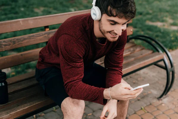 Deportista sonriente usando teléfonos inteligentes y auriculares en el banco en el parque - foto de stock