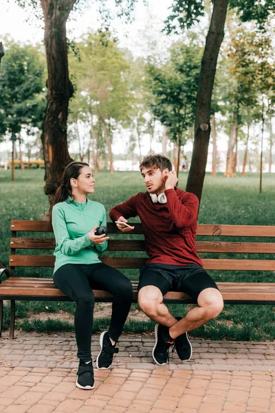 Улыбающаяся спортсменка держит спортивную бутылку возле бойфренда в наушниках со смартфоном на скамейке в парке — стоковое фото