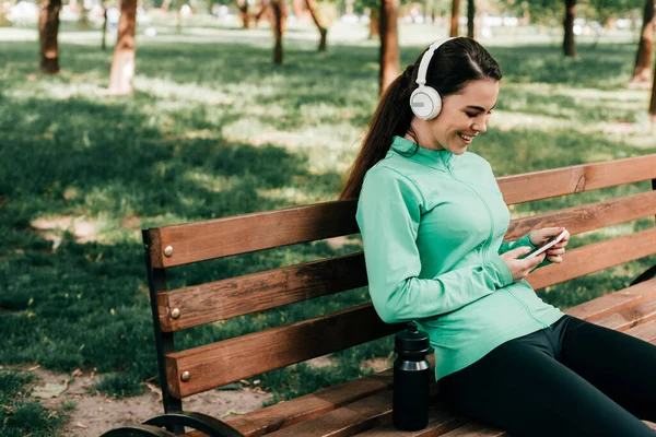 Sportiva sorridente in cuffia utilizzando smartphone vicino a bottiglia sportiva su panchina nel parco — Foto stock