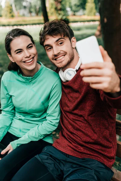 Выборочный фокус улыбающегося спортсмена, делающего селфи со смартфоном рядом с девушкой на скамейке в парке — стоковое фото