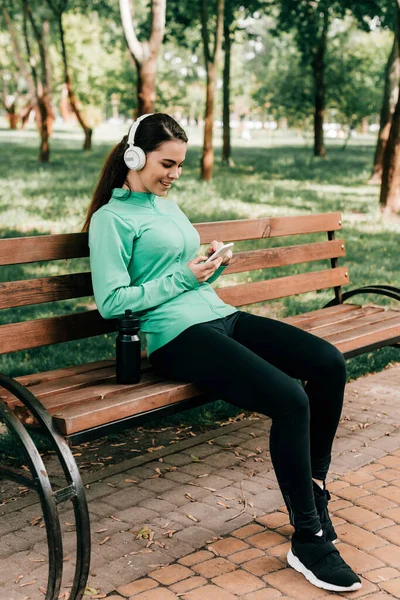 Sportiva sorridente in cuffia utilizzando smartphone vicino a bottiglia sportiva su panchina nel parco — Foto stock