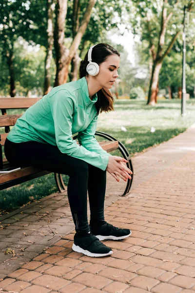 Vista lateral de la joven deportista en auriculares descansando en el banco en el parque - foto de stock