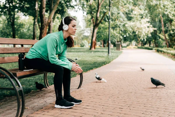 Side view of sportswoman in headphones sitting near sports bottle and smartphone on bench in park — Stock Photo