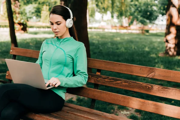 Femme dans les écouteurs en utilisant un ordinateur portable tout en étant assis sur le banc dans le parc — Photo de stock