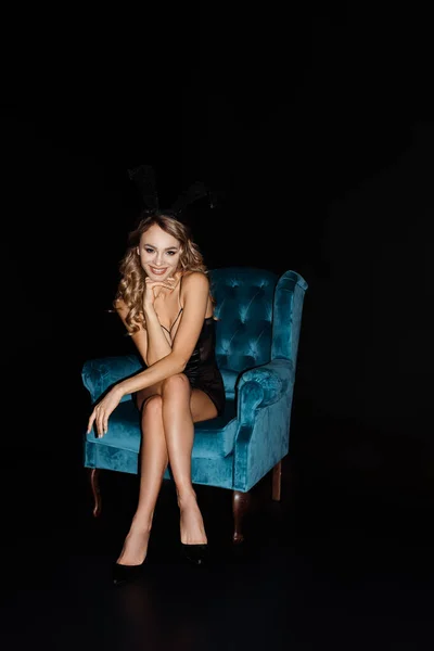 Mujer sexy en orejas de conejo sonriendo a la cámara mientras está sentado en sillón azul aislado en negro - foto de stock