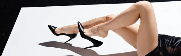 Panoramic crop of seductive woman in peignoir and heels lying on white surface on black background — Stock Photo
