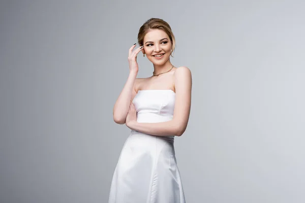 Mariée gaie en robe de mariée touchant les cheveux isolés sur gris — Photo de stock