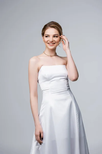 Happy bride in white wedding dress touching hair isolated on grey — Stock Photo