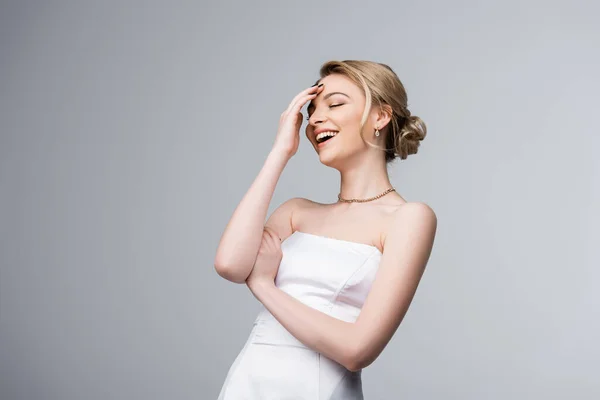 Novia feliz en vestido de novia blanco riendo mientras toca la frente aislada en gris - foto de stock