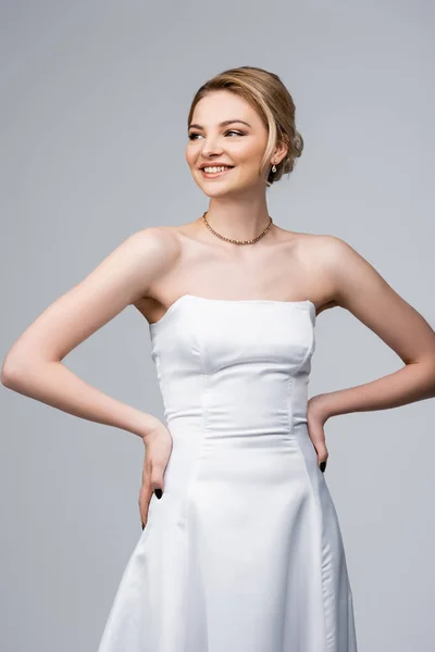 Atractiva novia en vestido de novia blanco sonriendo y de pie con las manos en las caderas aisladas en gris - foto de stock