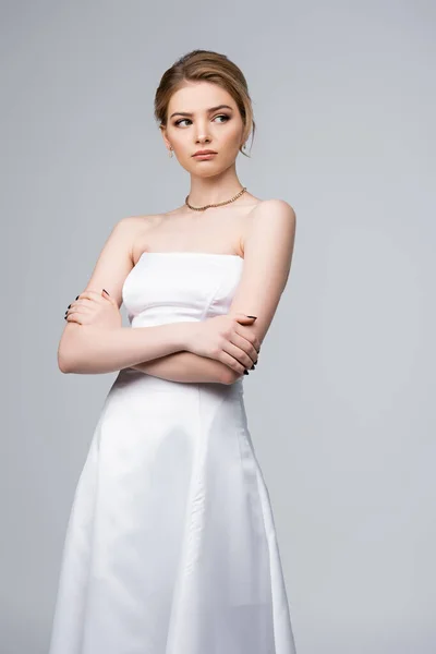 Novia disgustada en vestido de novia blanco de pie con brazos cruzados aislados en gris - foto de stock