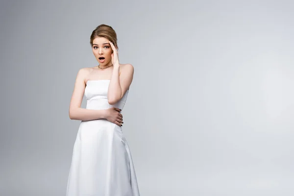Novia conmocionada en vestido de novia mirando a la cámara y tocando la cara aislada en gris - foto de stock