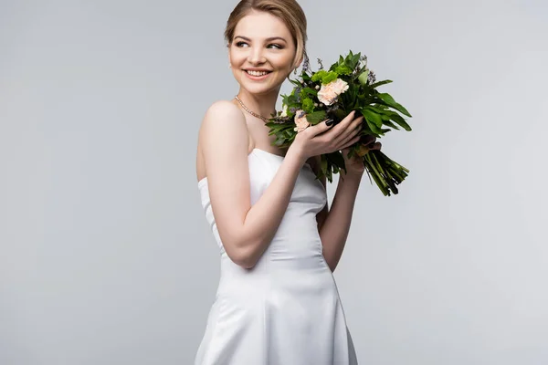 Novia alegre en vestido blanco sosteniendo flores de boda aisladas en gris - foto de stock