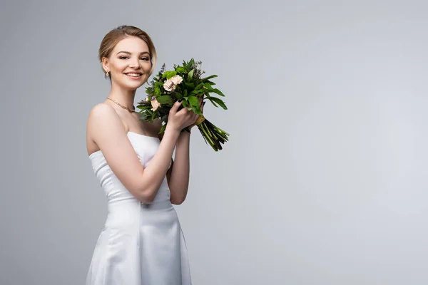 Mariée positive en robe blanche tenant des fleurs de mariage isolé sur gris — Photo de stock
