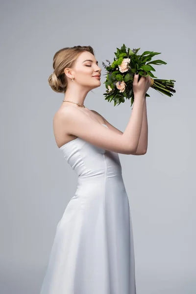 Positive bride in white dress smelling wedding flowers isolated on grey — Stock Photo