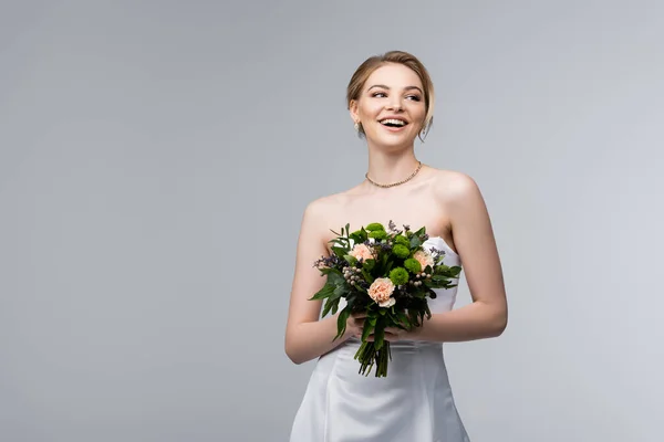 Fille positive en robe blanche tenant des fleurs de mariage isolé sur gris — Photo de stock