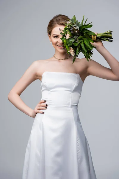 Positive girl in white dress holding wedding flowers near face and standing with hand on hip isolated on grey — Stock Photo