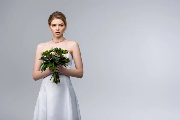 Noiva surpresa no vestido de casamento branco segurando flores isoladas no cinza — Fotografia de Stock