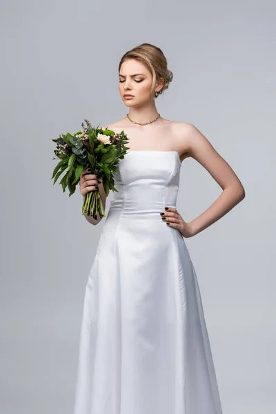 Atractiva novia en vestido blanco mirando flores de boda aisladas en gris - foto de stock