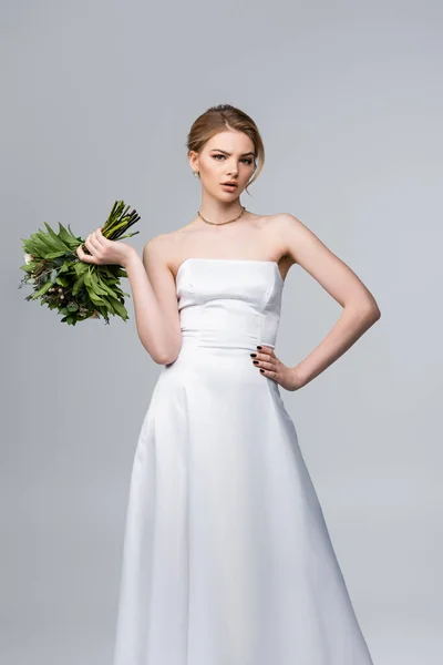 Jolie mariée en robe blanche tenant des fleurs de mariage et debout avec la main sur la hanche isolé sur gris — Photo de stock