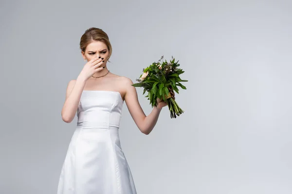 Bride in white wedding dress touching nose while holding smelly flowers isolated on grey — Stock Photo